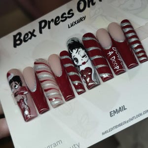 Puede incluir: Un juego de uñas postizas largas de punta cuadrada con una paleta de colores rojo oscuro y plateado. Los diseños incluyen un personaje de dibujos animados, remolinos de purpurina plateada, puntas rojas y el texto Boop Oop A Doop.