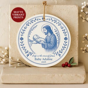May include: Round, white ornament with a blue illustration of Jesus holding a baby, surrounded by a floral border. The text "Safe in the arms of Jesus" and "Baby Adaline 2025" are printed on the ornament. A gold string is attached for hanging.