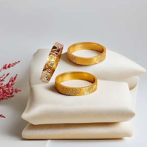 May include: Three gold rings with floral designs. The ring on the left has a floral design, the middle ring has the text "Love You Mom" engraved on it, and the ring on the right has a floral design.