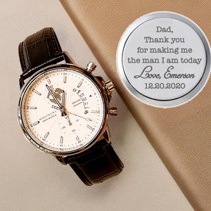 May include: A silver round engraved keepsake with the text "Dad, Thank you for making me the man I am today Love, Emerson 12.20.2020" and a brown leather wristwatch with a white face and gold accents.