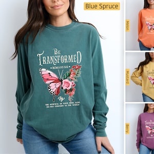 Può includere: Una t-shirt a maniche lunghe con un design a farfalla e fiori. La t-shirt ha il testo "Be Transformed" con il riferimento biblico "Romani 12:2" e la frase "Anche per il rinnovamento della tua mente, non conformarti al mondo". La t-shirt è disponibile in quattro colori: abete blu, melone, senape e bacca.