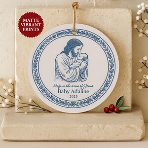 Puede incluir: Adorno redondo blanco con una ilustración azul de Jesús sosteniendo a un bebé, rodeado por un borde floral. El texto "Safe in the arms of Jesus Baby Adaline 2025" está impreso debajo. Una pegatina roja mate está en la esquina superior izquierda.