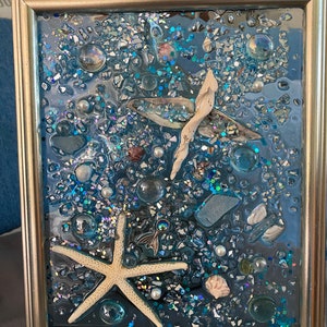 May include: A framed resin art piece with a blue background and various sea-themed elements, including a starfish, seashells, and glitter.