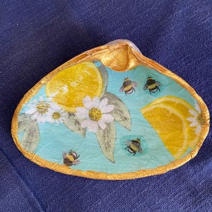 May include: A gold-rimmed seashell dish with a blue background and a lemon and bee design. The dish is decorated with yellow lemons, white daisies, and black and yellow bees.