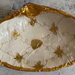 May include: A white seashell dish with a gold rim and a honeycomb pattern with bees. The dish is perfect for holding small items like jewelry or trinkets.