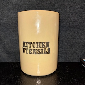 May include: A cylindrical beige ceramic kitchen utensil holder with a smooth, glossy finish. The words KITCHEN UTENSILS are printed in bold, black, capital letters on the front centre of the container. The item has a flat base and an open top.