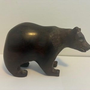 May include: A dark brown wooden bear figurine. The bear is carved with visible texture and detail, showcasing its fur and form. The figurine is posed in a quadruped stance, with its head slightly raised.