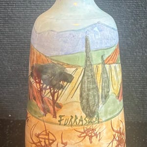 May include: A ceramic bottle with a painted landscape design. The bottle features a light blue top, a brown rim, and a landscape scene with trees, fields, and mountains. The word "FURRASTAS" is written on the bottle.