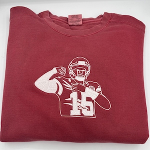 Puede incluir: Una sudadera color granate con una imagen bordada en blanco de un jugador de fútbol americano con casco y camiseta con el número 15.