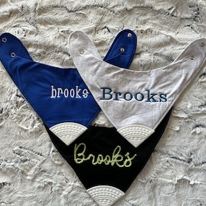 May include: Three baby bibs with the name "Brooks" embroidered on them. The bibs are in blue, gray, and black with white trim.