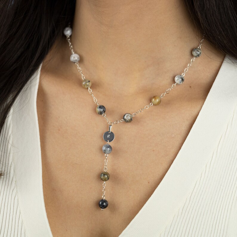 May include: A silver chain necklace with a silver pendant featuring the letter "E". The necklace has a layered design with a shorter strand of beads and a longer strand of beads. The beads are a mix of black, white, and brown.