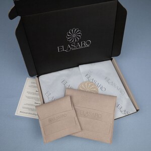May include: A black box with the logo "ELASARO" printed on the top. The box is open and contains two small beige pouches with the same logo. The pouches are on top of white tissue paper.
