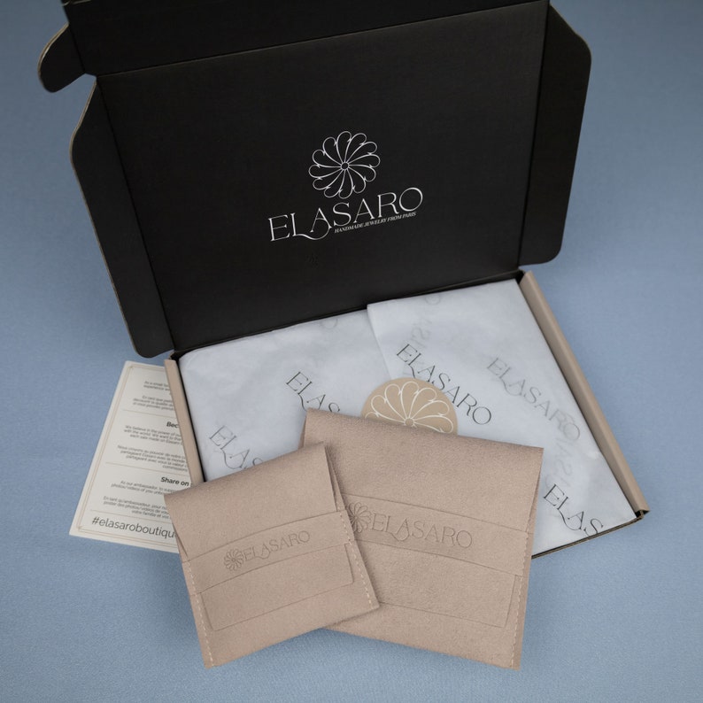 May include: A black box with the logo "ELASARO" printed on the top. The box is open and contains two small beige pouches with the same logo. The pouches are on top of white tissue paper.