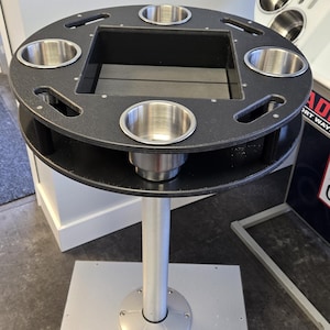 May include: A black metal table with a circular top featuring six stainless steel cup holders. The table has a central recessed area and is supported by a silver pole and a square base.