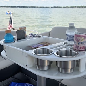 May include: A white boat table with stainless steel cup holders and a central storage area. The table holds a bottle of water, a can of soda, and other items. The boat is on a lake with a boat and people in the background.