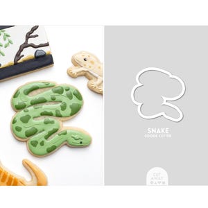 May include: Cookies decorated as a green snake with darker green spots, a lizard, and a landscape. A white cookie cutter in the shape of a snake is also shown. The text "SNAKE COOKIE CUTTER" is visible.