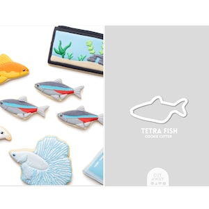 May include: An assortment of decorated fish-shaped cookies, including a goldfish, tetra fish, and a betta fish. The cookies are arranged with a cookie cutter outline and a miniature aquarium cookie. The text "TETRA FISH COOKIE CUTTER" is visible.