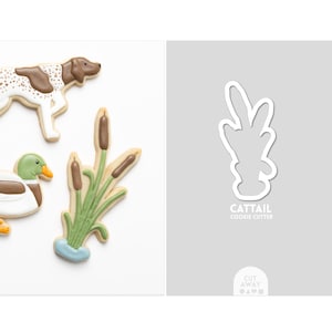 May include: Image features decorated cookies: a dog, a duck, and cattails. The cattail cookie is next to a cookie cutter with the text "CATTAIL COOKIE CUTTER". The cookies are on a white background.