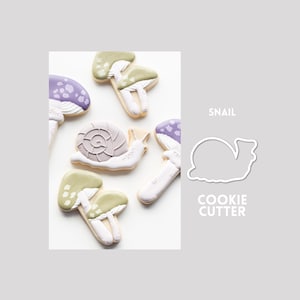 May include: A snail cookie cutter and several decorated cookies shaped like snails and mushrooms. The cookies feature white, sage green, and lavender icing with detailed patterns. The text SNAIL COOKIE CUTTER is printed in white on the right side.