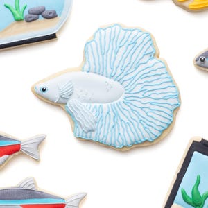 May include: Assortment of fish-shaped cookies, including a white betta fish with blue accents, a goldfish, and other colorful fish designs. The cookies are decorated with intricate details and placed on a white background. The cookies are a fun, whimsical treat.