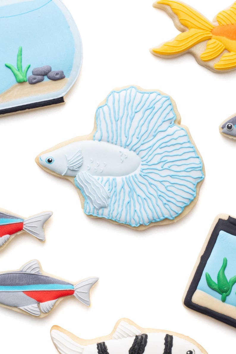 May include: Assortment of fish-shaped cookies, including a white betta fish with blue accents, a goldfish, and other colorful fish designs. The cookies are decorated with intricate details and placed on a white background. The cookies are a fun, whimsical treat.