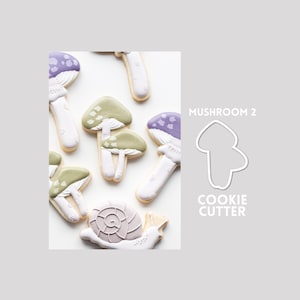 May include: A collection of decorated sugar cookies shaped like mushrooms and a snail, alongside a white outline of a MUSHROOM 2 COOKIE CUTTER. The cookies feature white stems with purple or sage green caps decorated with white spots.