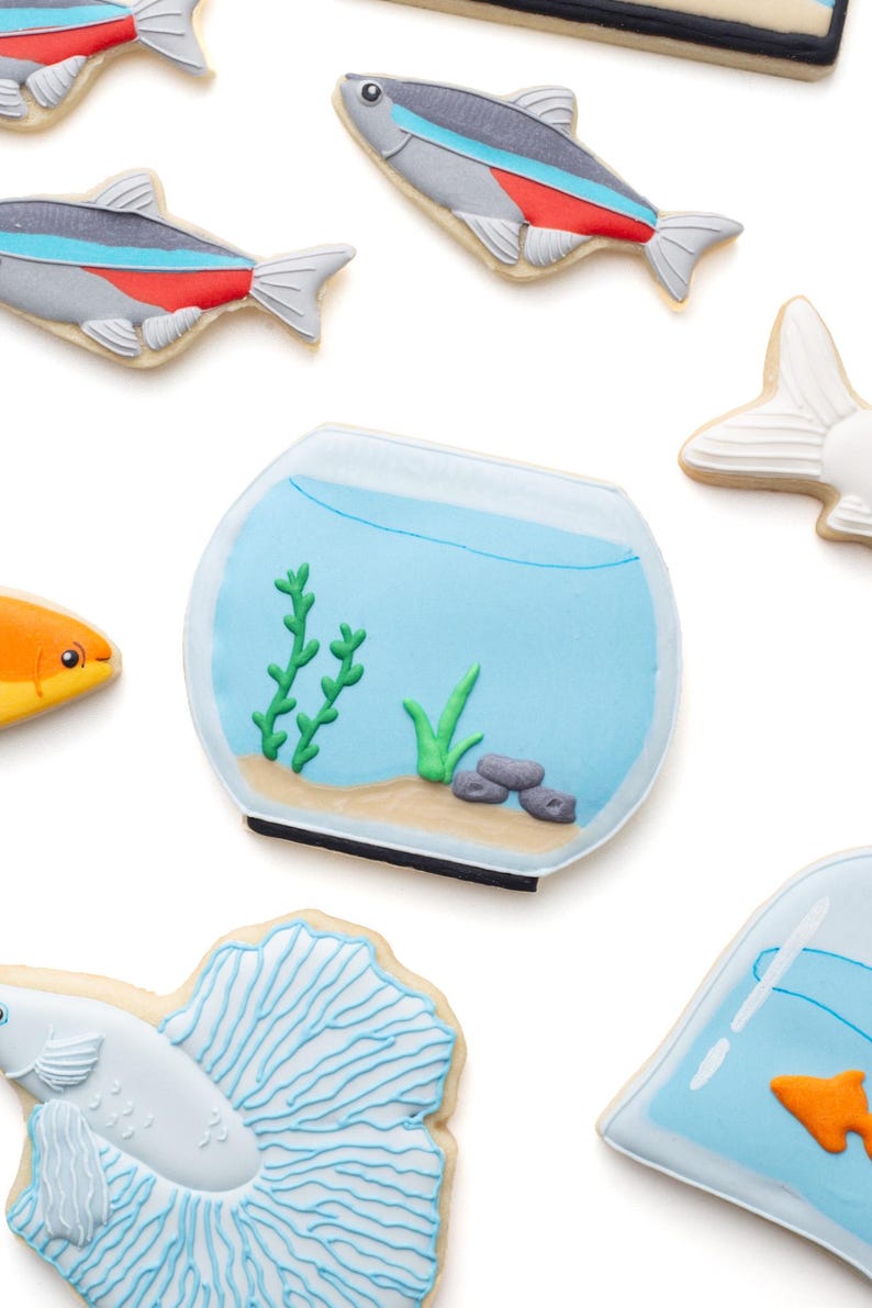 May include: Assortment of fish-themed cookies, including a fishbowl with aquatic plants and several fish cookies. The fish cookies feature various designs, including a neon tetra with red and blue stripes, and a betta fish with a detailed tail.