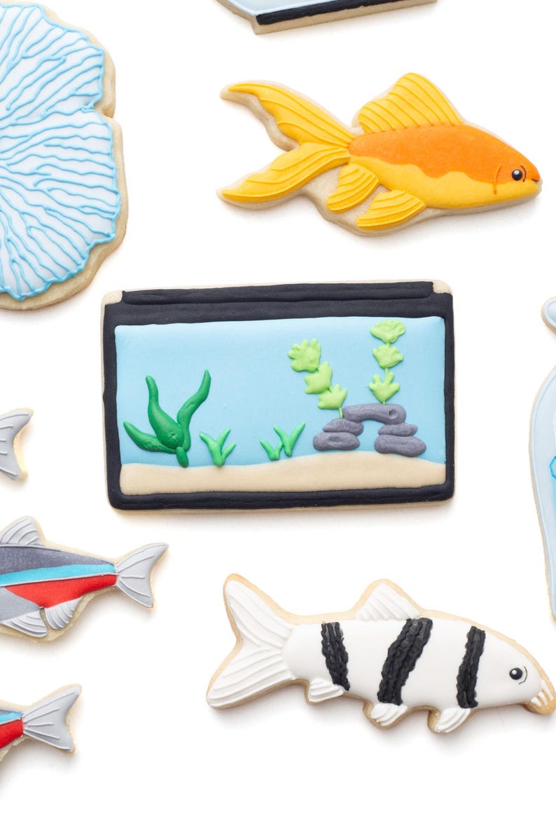 May include: Assortment of decorated sugar cookies in the shape of aquatic life. Includes a goldfish, a fish tank with plants, and various fish designs. The cookies are decorated with bright colors and intricate details, perfect for an ocean-themed event.
