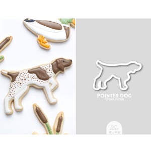 May include: An image features a pointer dog cookie and a cookie cutter. The cookie is white with brown spots and a brown head. The cookie cutter is a white outline of a pointer dog. The text "POINTER DOG COOKIE CUTTER" is below the cutter.