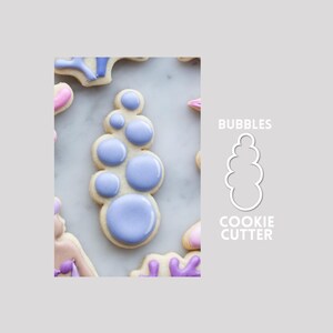 Op de afbeelding: Een koekje in de vorm van een verticale tros bellen, versierd met glad, glanzend lichtpaars glazuur. Naast het koekje staat een witte omtrektekening van dezelfde vorm met de tekst BUBBLES COOKIE CUTTER in het wit gedrukt.