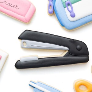 May include: A collection of school supply-shaped sugar cookies including a black and silver stapler, a pink cookie with the word Eraser, a blue and white pencil case with a green zipper, and a blue correction tape dispenser with a white applicator.