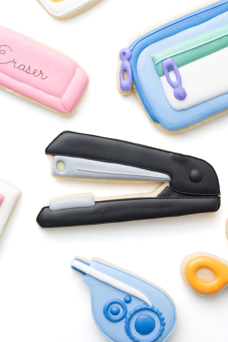 May include: A collection of school supply-shaped sugar cookies including a black and silver stapler, a pink cookie with the word Eraser, a blue and white pencil case with a green zipper, and a blue correction tape dispenser with a white applicator.
