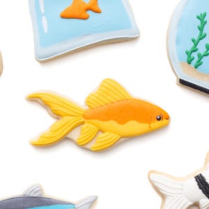 May include: Assortment of fish-shaped cookies, including a goldfish, a fish in a bag, and other fish designs. The goldfish cookie is orange and yellow, with detailed fins. The cookies are on a white background.