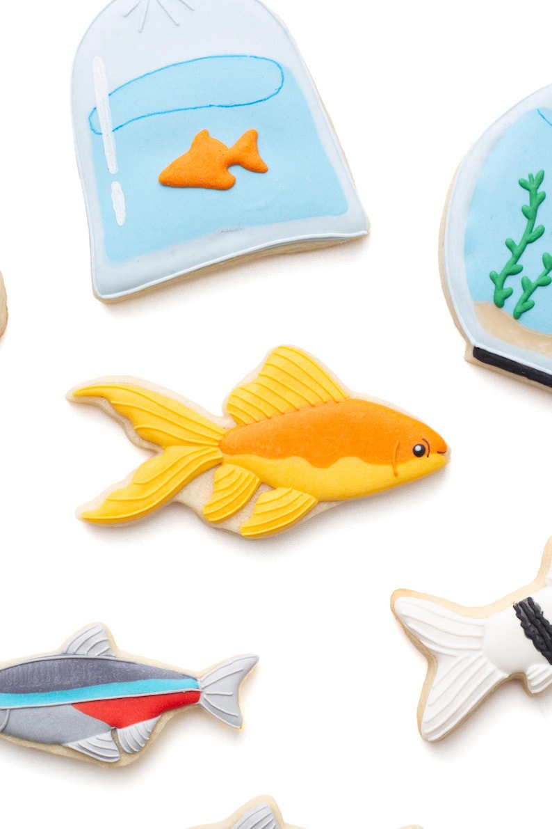 May include: Assortment of fish-shaped cookies, including a goldfish, a fish in a bag, and other fish designs. The goldfish cookie is orange and yellow, with detailed fins. The cookies are on a white background.