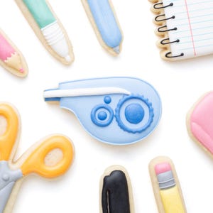 May include: A collection of school supply shaped sugar cookies including a spiral notebook, a correction tape dispenser, scissors, a black marker, a yellow pencil with a pink eraser, and various pens in shades of light blue, mint green, and pink.