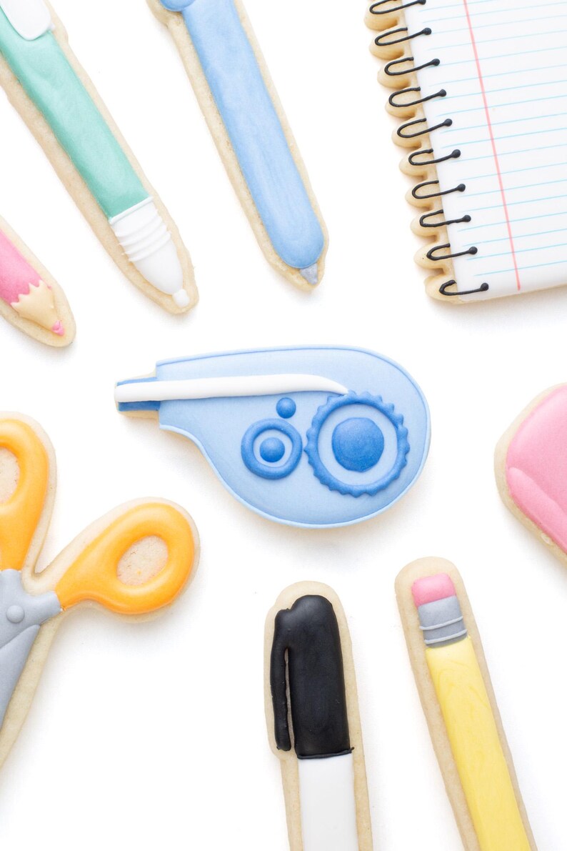 May include: A collection of school supply shaped sugar cookies including a spiral notebook, a correction tape dispenser, scissors, a black marker, a yellow pencil with a pink eraser, and various pens in shades of light blue, mint green, and pink.