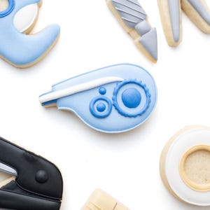 May include: A collection of office supply shaped sugar cookies including a light blue tape dispenser, a gray craft knife, orange and gray scissors, a blue correction tape, a black stapler, a beige ruler, and a white roll of adhesive tape.
