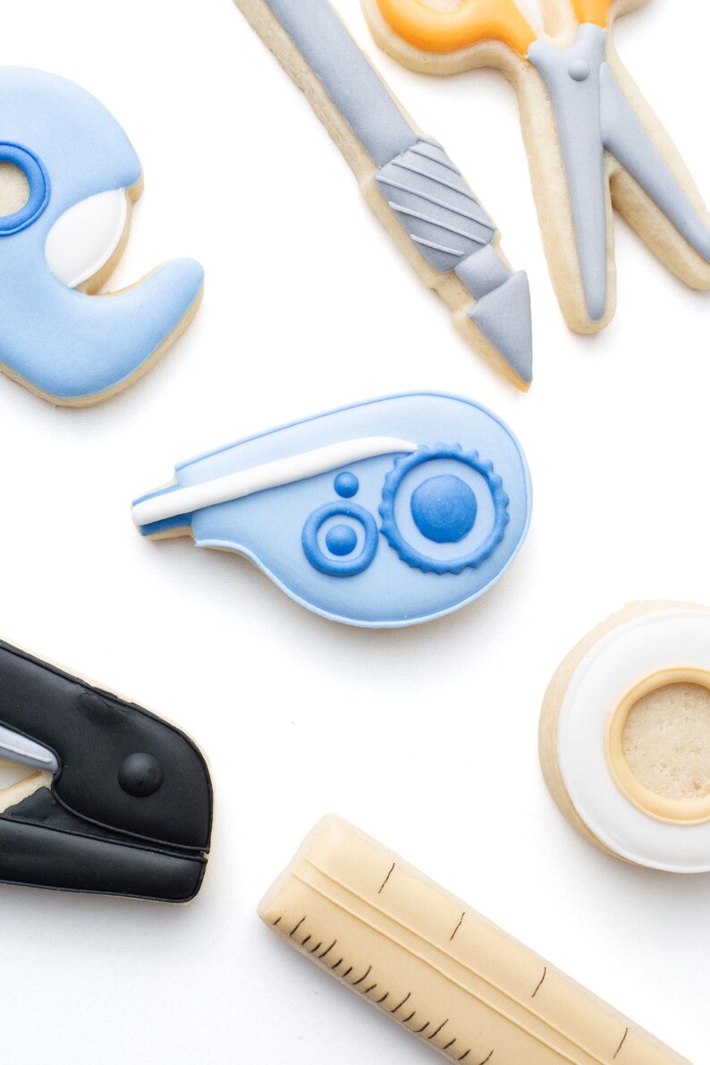 May include: A collection of office supply shaped sugar cookies including a light blue tape dispenser, a gray craft knife, orange and gray scissors, a blue correction tape, a black stapler, a beige ruler, and a white roll of adhesive tape.