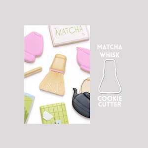 Peut inclure: Une collection de biscuits décorés comprenant un fouet à matcha beige, un bol rose, une théière noire et un biscuit rectangulaire avec le texte MATCHA. À côté des biscuits se trouve le contour blanc d'un emporte-pièce avec le texte MATCHA WHISK COOKIE CUTTER.