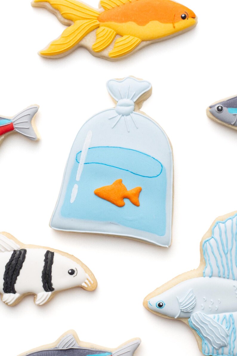 May include: Assortment of fish-shaped cookies, including a goldfish, a fish in a bag, and other colorful fish designs. The cookies are decorated with orange, yellow, blue, black, and white icing. A fun treat for an aquatic-themed party.