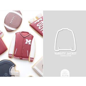 May include: Decorated cookies in the shape of American football-themed items, including a maroon and white varsity jacket with the letter "M", a football, and a helmet. A cookie cutter outline of a varsity jacket is also shown.