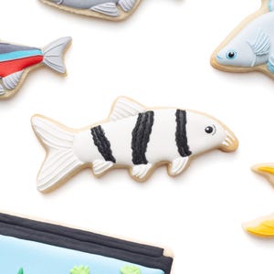May include: Assortment of fish-shaped cookies with detailed icing. The cookies feature various designs, including a black and white striped fish, and fish with red, blue, and gray accents. The cookies are on a white background.