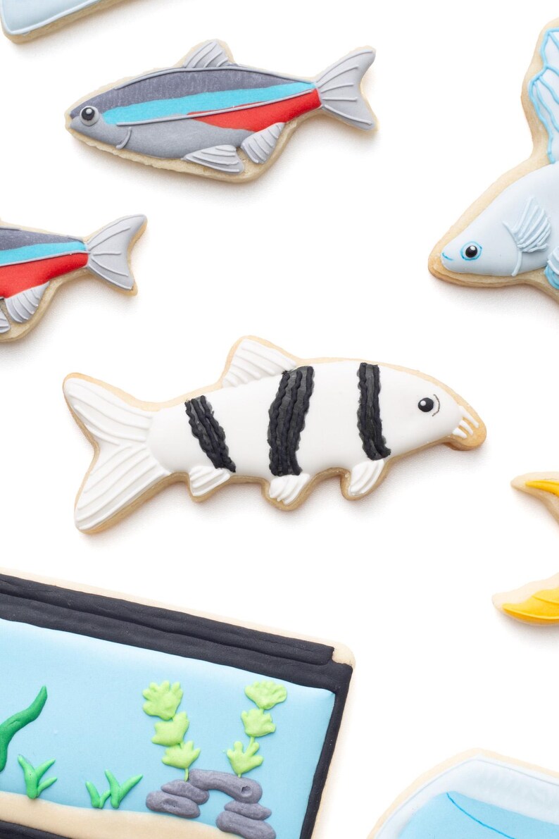 May include: Assortment of fish-shaped cookies with detailed icing. The cookies feature various designs, including a black and white striped fish, and fish with red, blue, and gray accents. The cookies are on a white background.