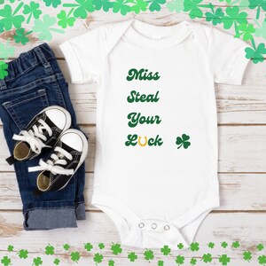 Pode incluir: Macacão branco para bebê com texto verde que diz "Miss Steal Your Luck" com uma ferradura dourada e um trevo verde.