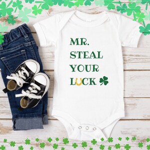 Pode incluir: Macacão branco para bebê com texto verde que diz "MR. STEAL YOUR LUCK" com uma ferradura dourada e um trevo.