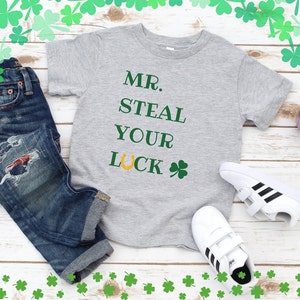 Pode incluir: Uma camiseta cinza com o texto "MR. STEAL YOUR LUCK" em verde e dourado. A camiseta é combinada com jeans azul e tênis brancos com listras pretas.