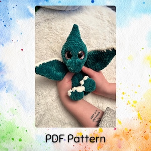May include: A green crocheted dinosaur toy with white accents and large, glittery eyes. The toy is being held by a person's hands. The image includes the text "PDF Pattern".