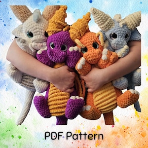 May include: A collection of handmade crochet dragons in various colors, including purple, orange, blue, and cream. Each dragon has large eyes and detailed features. The image includes the text "PDF Pattern".
