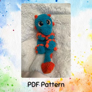 Op de afbeelding: Een handgemaakt gehaakt draakspeelgoed in turquoise en oranje met grote, glanzende ogen. De draak heeft een lang, slank lichaam en ligt op een zacht, grijs oppervlak. De tekst "PDF Pattern" staat onderaan de afbeelding.