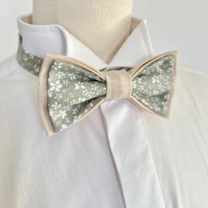 Peut inclure: Un nœud papillon à motif floral vert clair avec des fleurs blanches. Il présente une bordure et un centre beiges, le tout sur une chemise blanche. Un accessoire élégant.